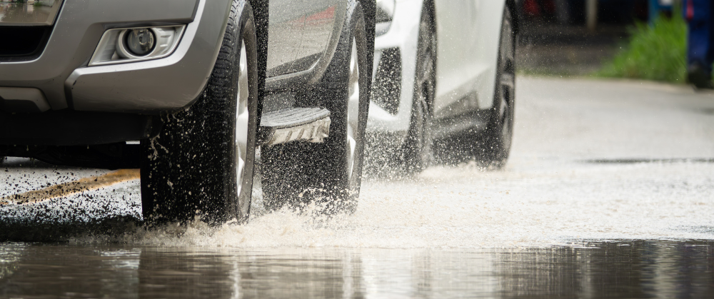 Fall Driving Hazards: How to Keep Your Car Safe on Slippery Roads. Close-up of a silver and white car driving through a flooded road, with water splashing from the tires.