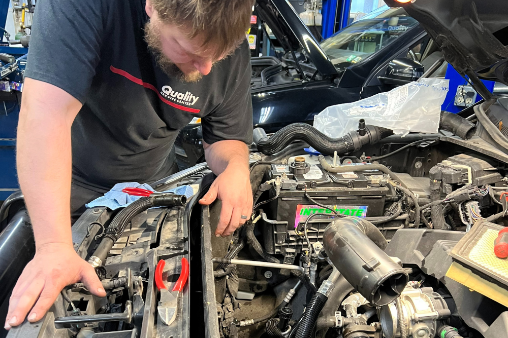 Car Maintenance in York, PA by Quality Service Center. Mechanic leaning over an open car engine, inspecting parts with tools and cables spread around the engine bay.